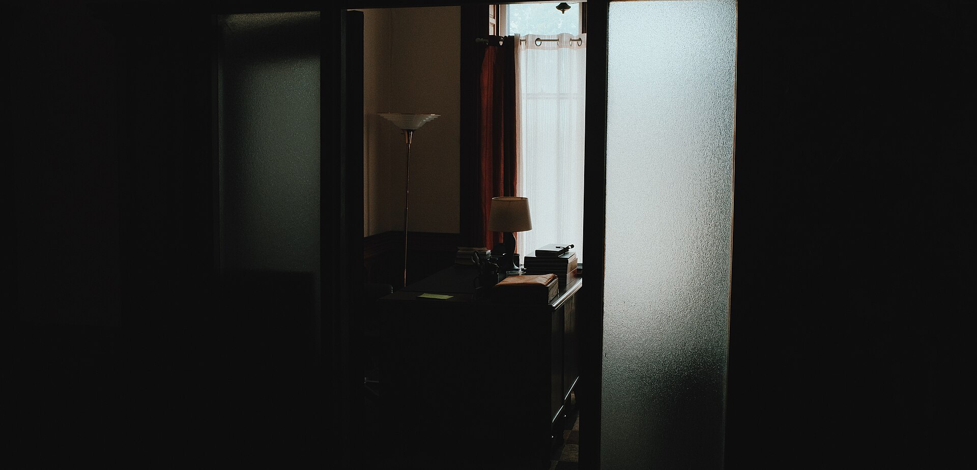 A dim interior seen through a part-open door: a desk, a shaded lamp and a curtained window in the room beyond, with no one present.