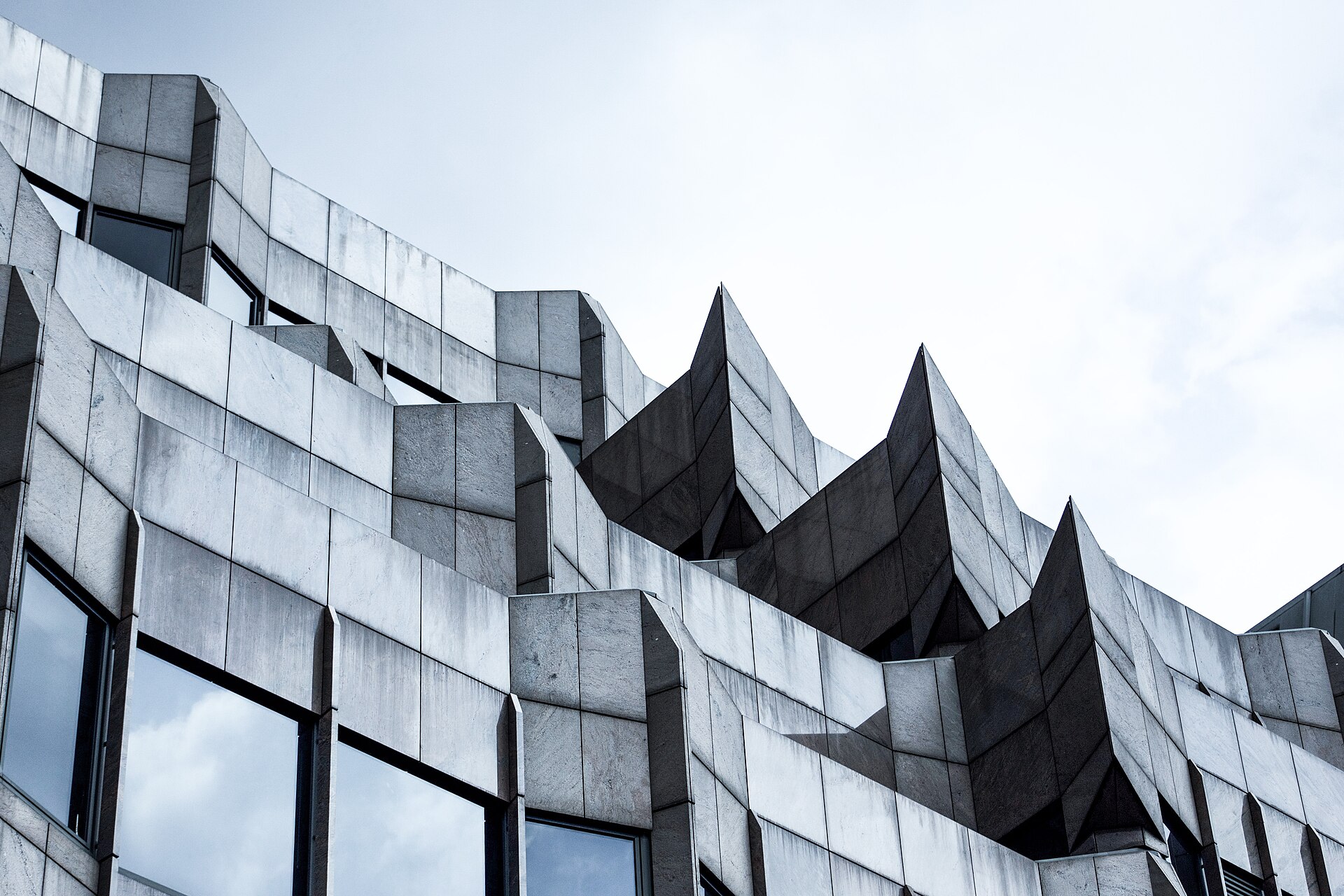 Angular precast cladding panels on a building corner, pale flat bays meeting a run of dark folded pleats against open sky.