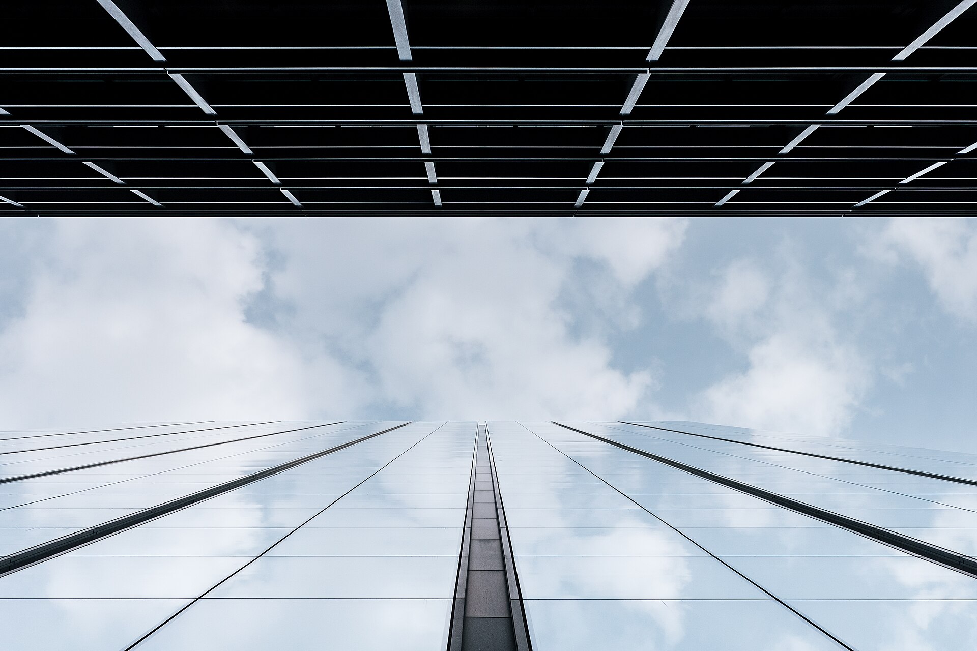 A glass office facade photographed from street level, its panels reflecting cloud, with a canopy of louvres overhead.
