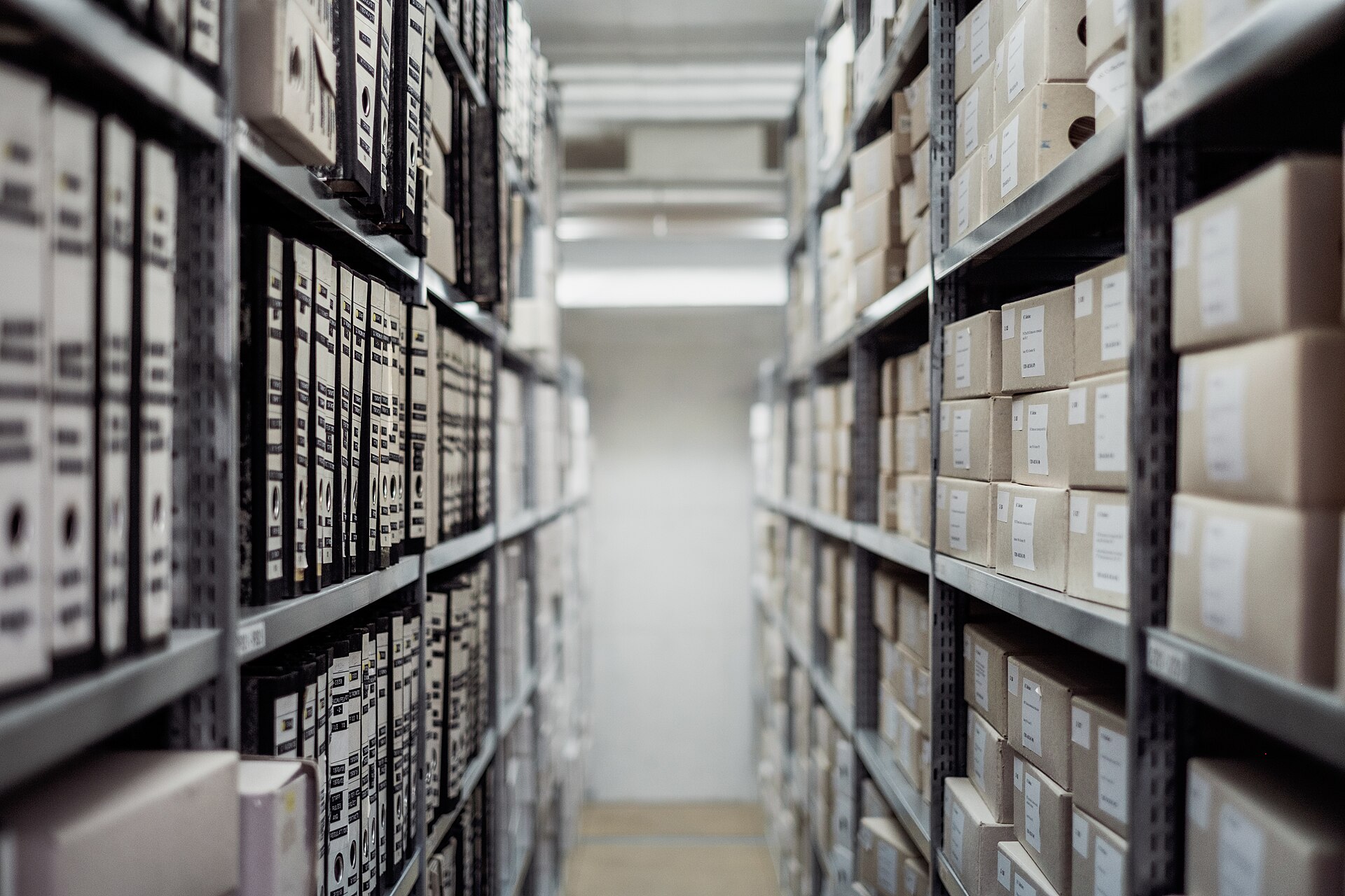 An aisle between two racks of archive shelving, stacked with labelled document boxes and ring binders.